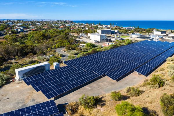 A large array of solar panels, sitting in front of Christies Beach wastewater Treatment Plant. Behind the Plant, to the left are trees and scattered housing, and to the right is the ocean. 
